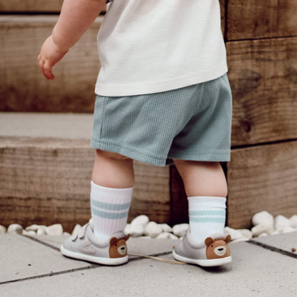 Kid's grey barefoot trainers with a brown bear face motif on the heel, pictured outdoors.