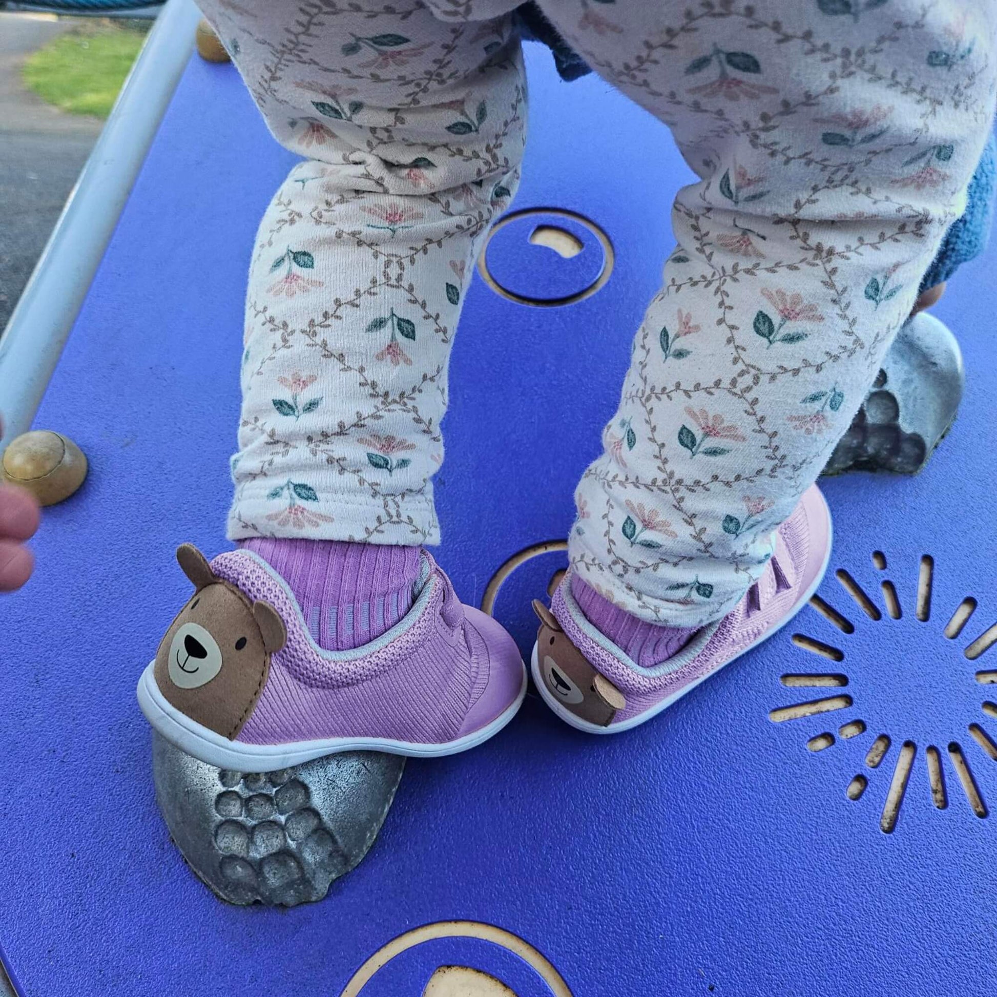 Kid's lilac barefoot trainers with a brown bear face design on the heel on an outdoor playground platform.