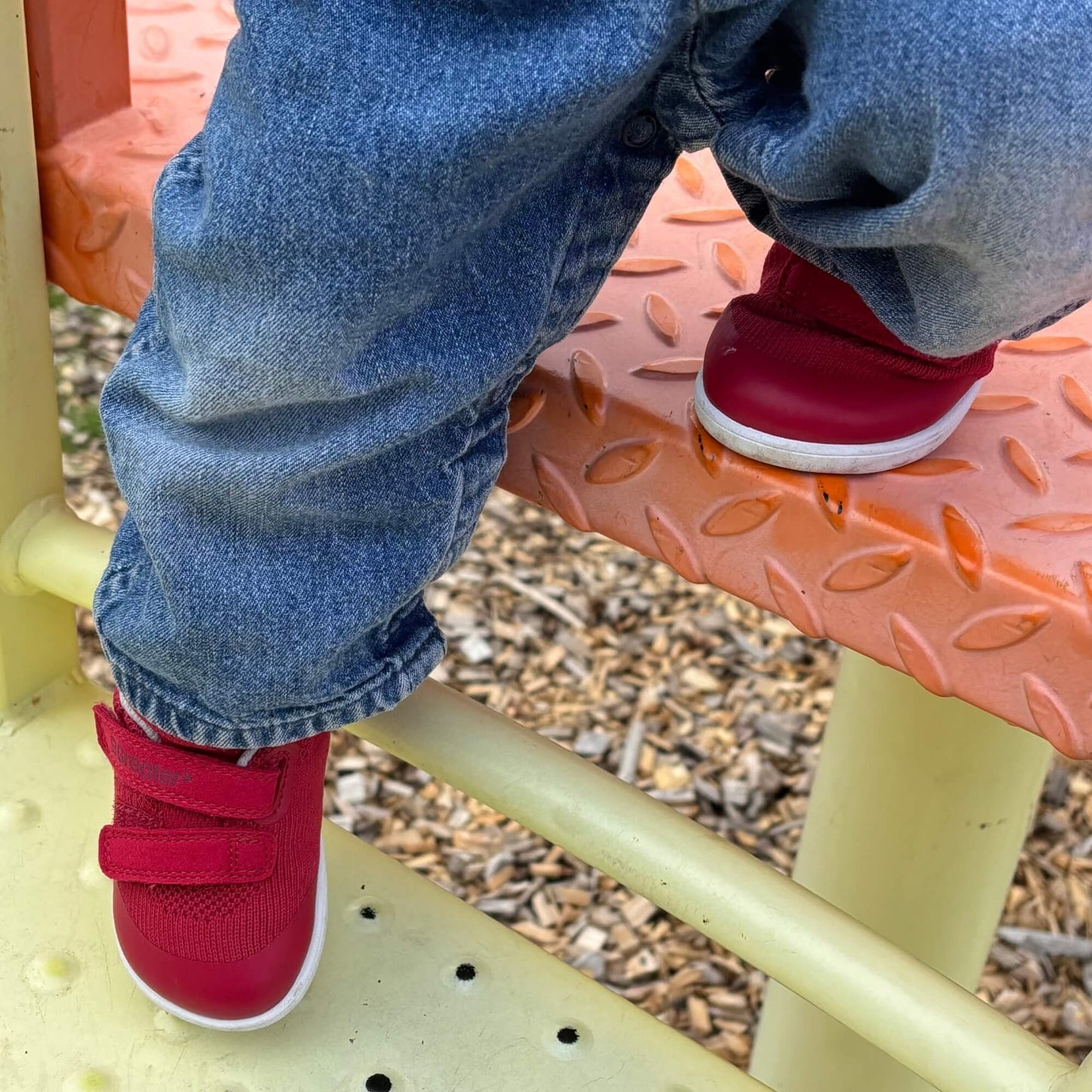 Kid's red barefoot trainers with hook-and-loop straps on an outdoor playground step.
