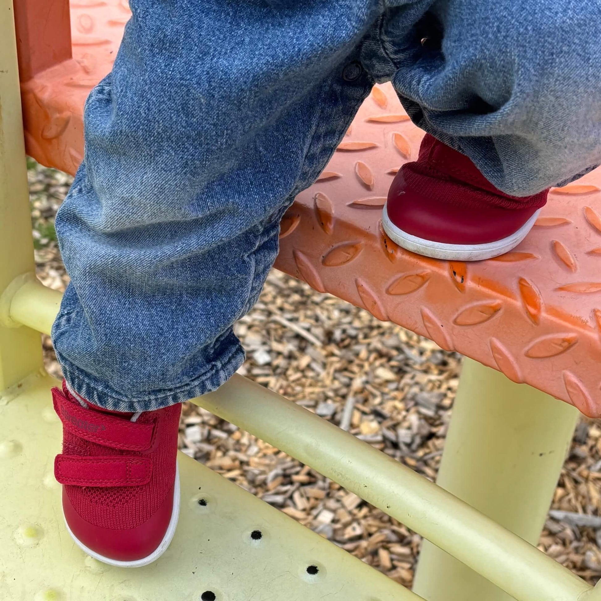 Kid's red barefoot trainers with hook-and-loop straps on an outdoor playground step.