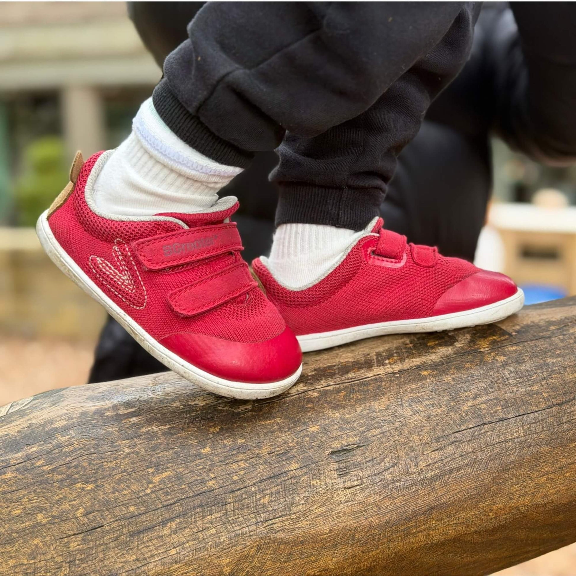 Kid's red barefoot trainers on an outdoor wooden log.