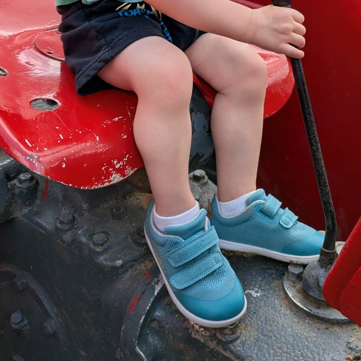 Kid wearing teal barefoot trainers sitting on an outdoor tractor.