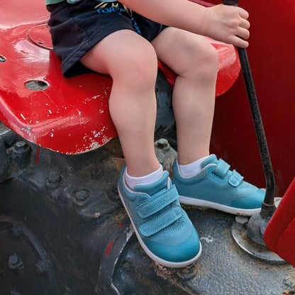 Kid wearing teal barefoot trainers sitting on an outdoor tractor.