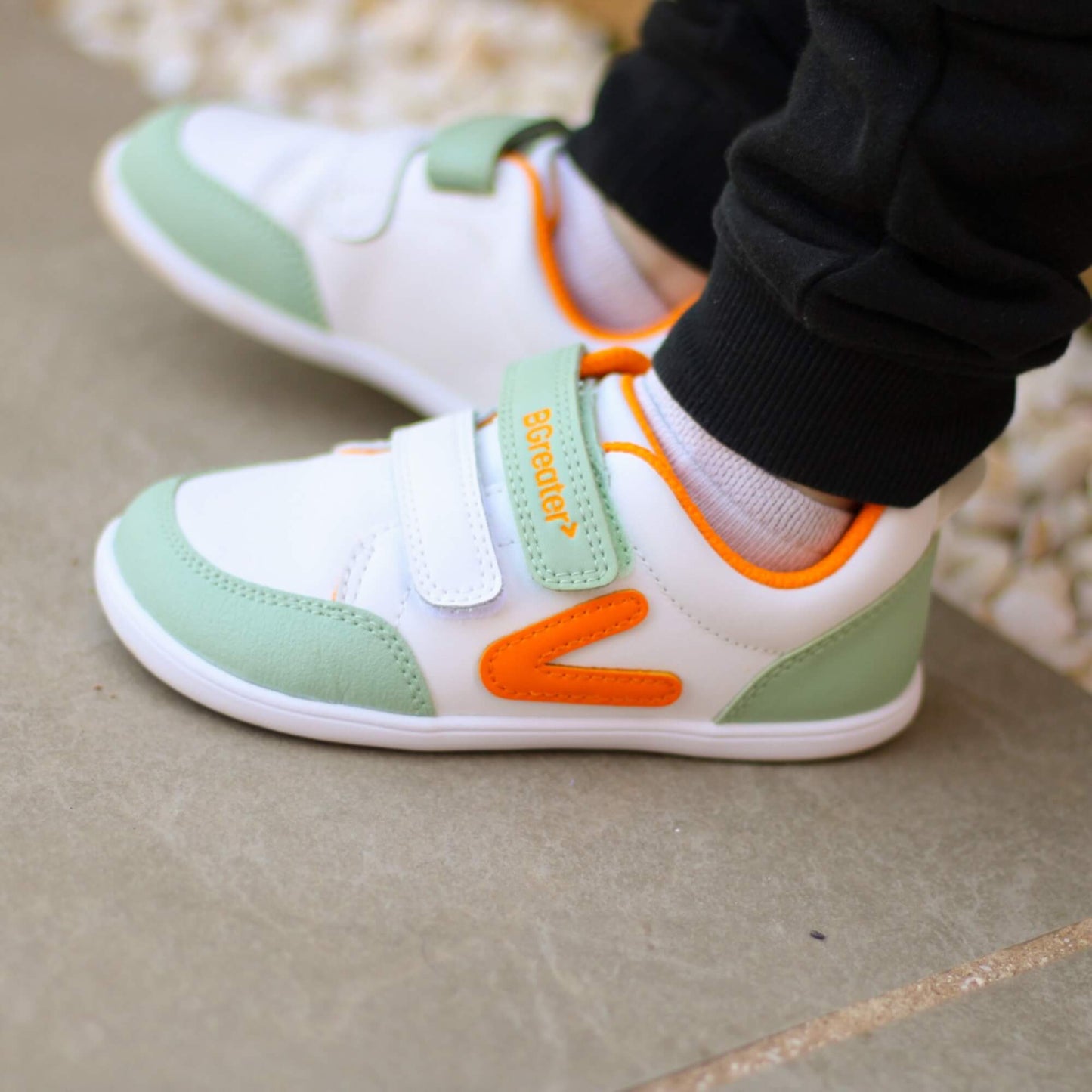 Kid's white, olive green and orange barefoot trainers on an outdoor stone patio.