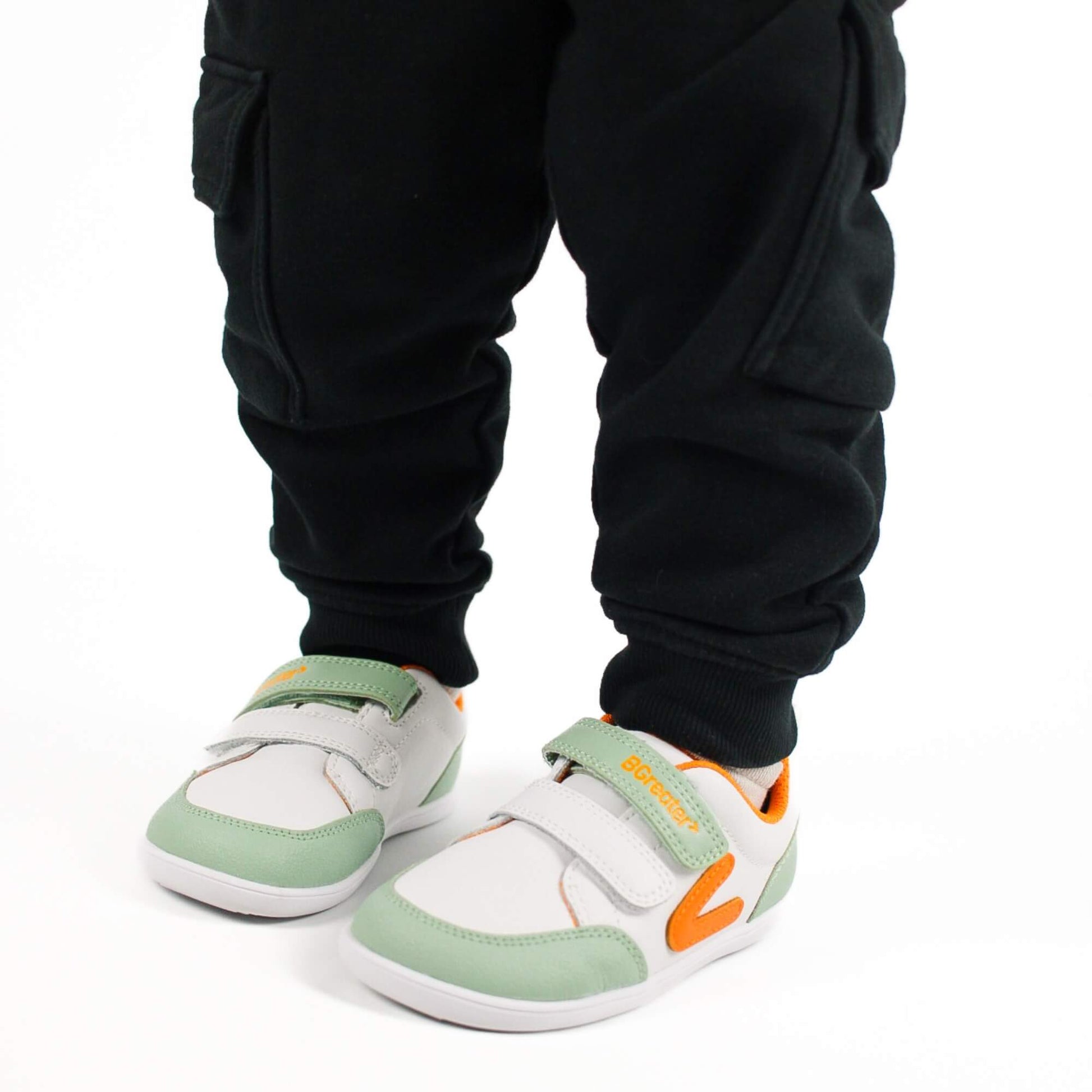 Close-up of a kid wearing white and olive green barefoot trainers with hook-and-loop straps on a white background.