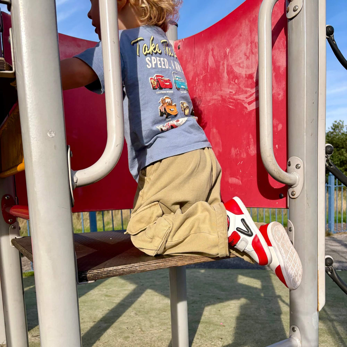 Kid wearing red and white leather barefoot trainers kneeling on an outdoor playground platform.