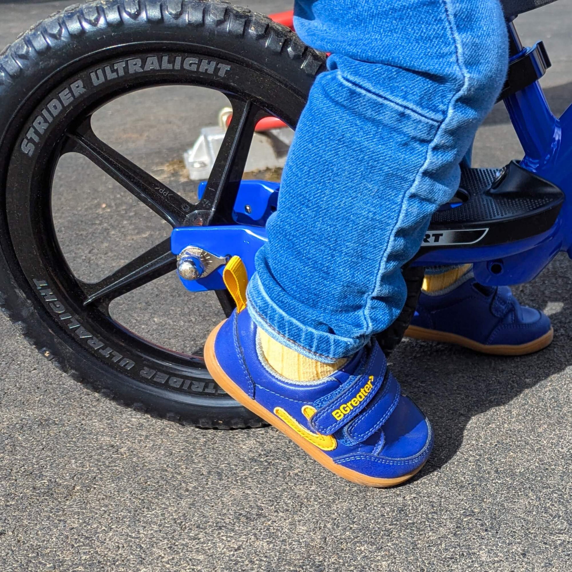 Child’s blue barefoot trainers with hook-and-loop straps and gum soles beside a balance bike wheel on outdoor pavement
