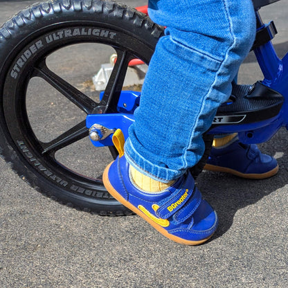 Child’s blue barefoot trainers with hook-and-loop straps and gum soles beside a balance bike wheel on outdoor pavement