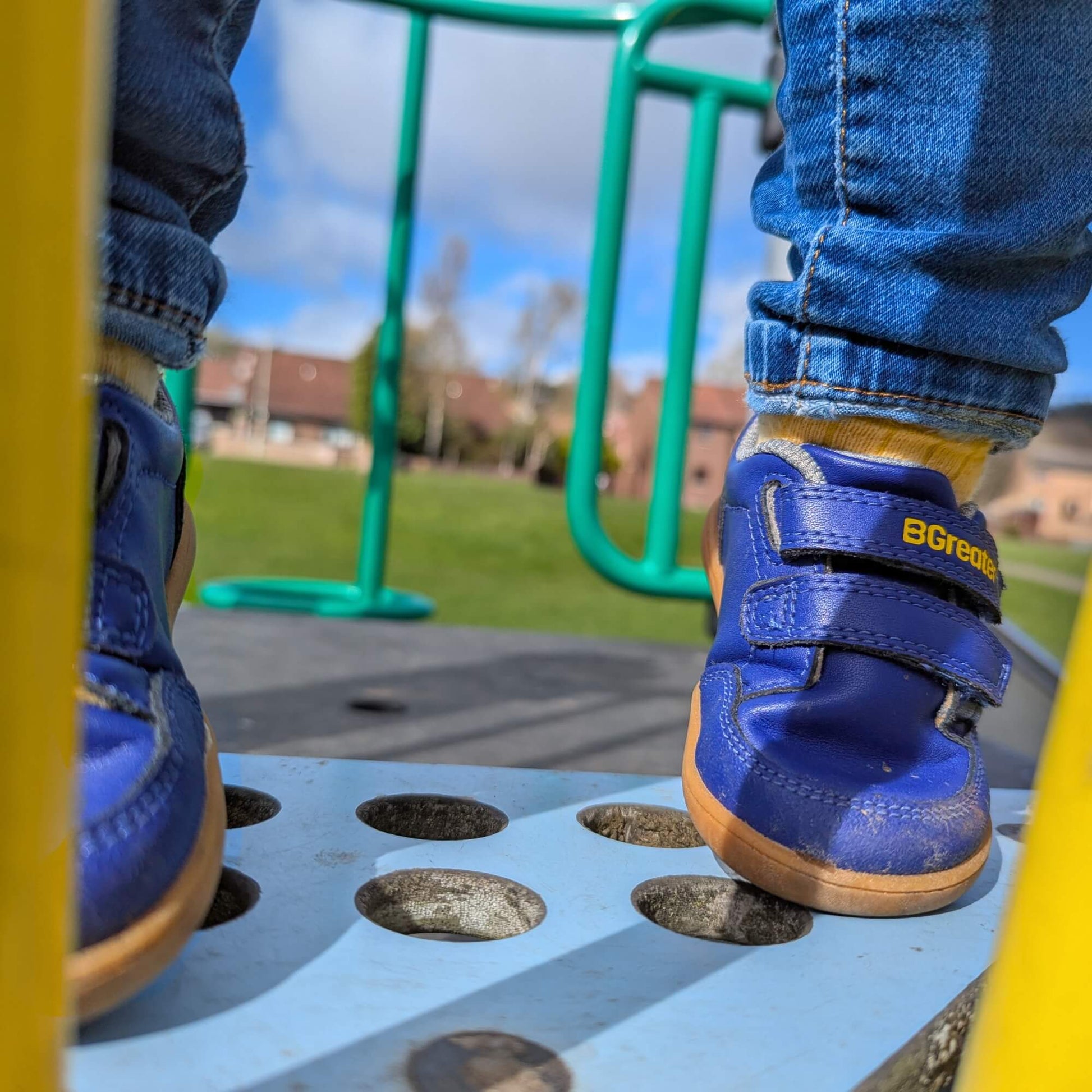Kid wearing blue barefoot trainers with double hook-and-loop straps and gum soles on a playground platform outdoors