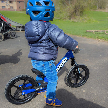 Kid wearing blue barefoot trainers with strap closure and gum soles while riding a balance bike in an outdoor park setting