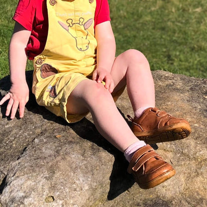 Kid wearing tan leather barefoot trainers with hook-and-loop straps outdoors.