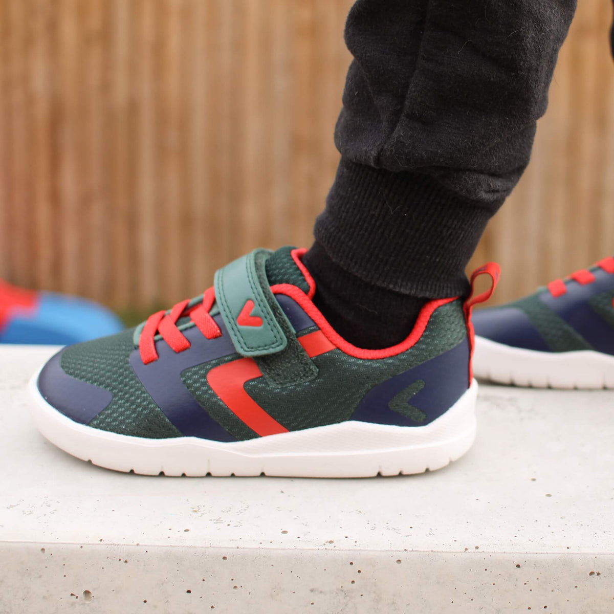 Side view of kid's green, navy and red barefoot trainers with a hook-and-loop strap outdoors.