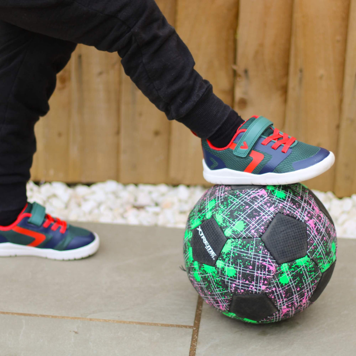 Kid wearing green and navy barefoot trainers with foot resting on a football outdoors.