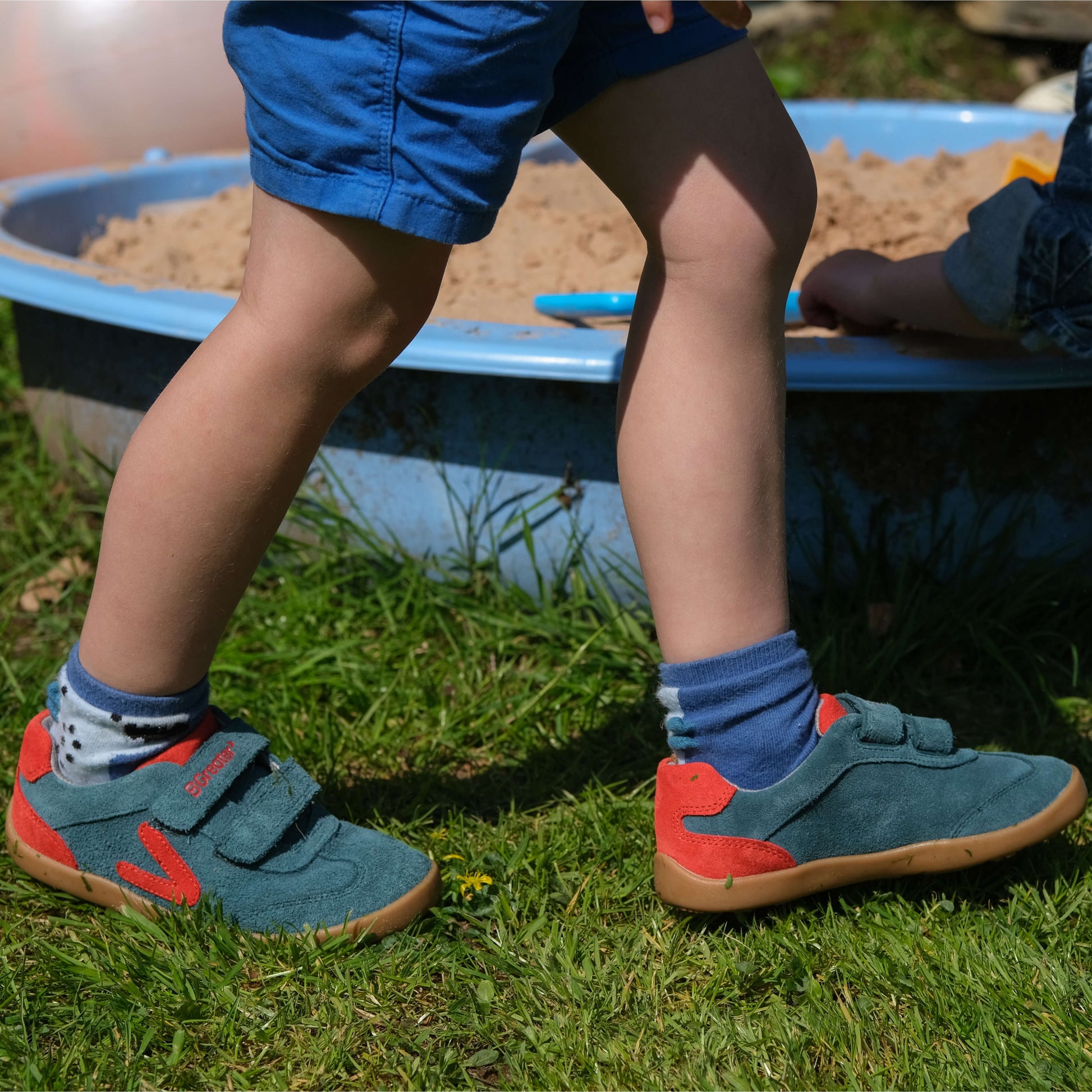 Child wearing green shoes with red soles standing on grass next to a sand pit.