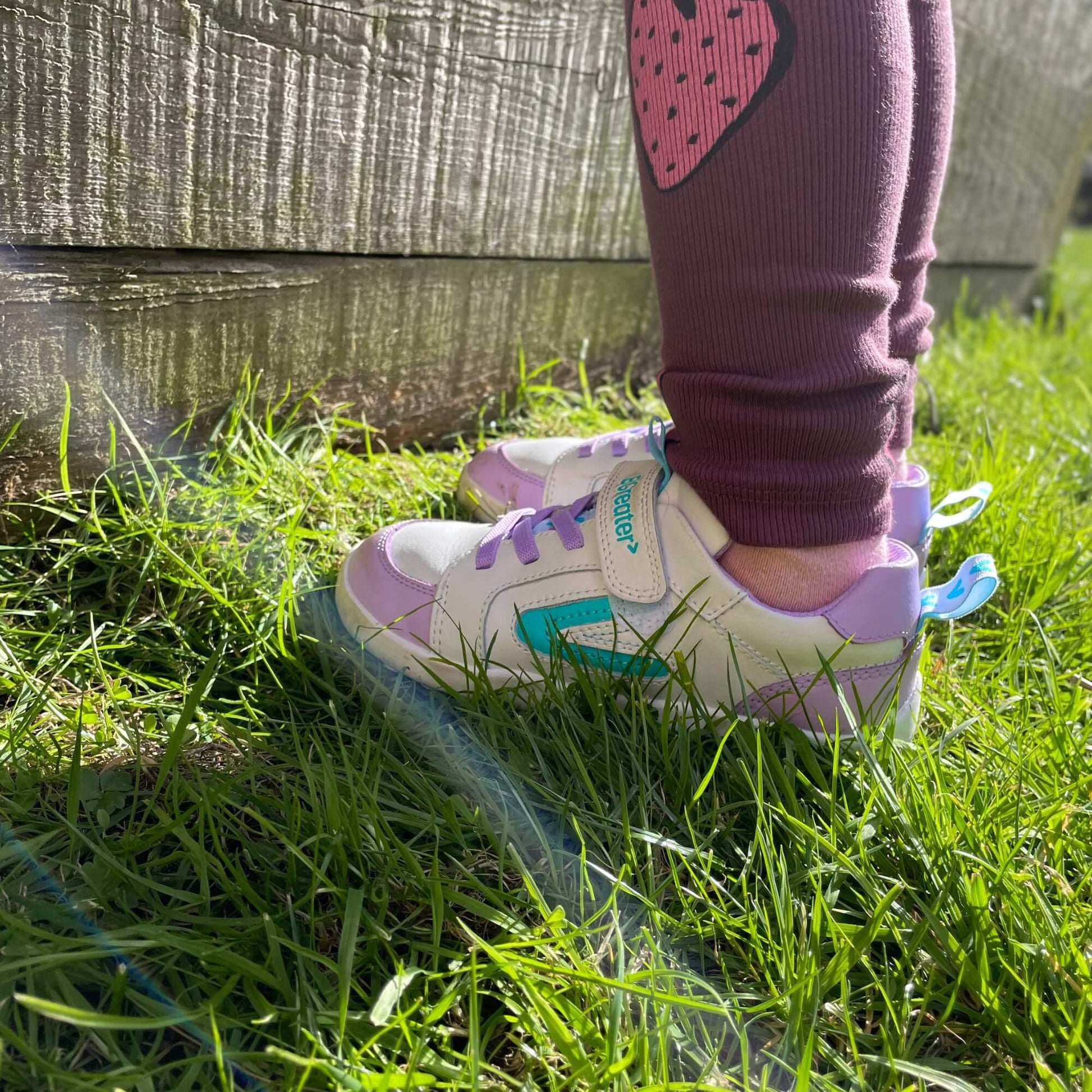 Kid’s white and lilac leather barefoot trainers with teal accents on outdoor grass.
