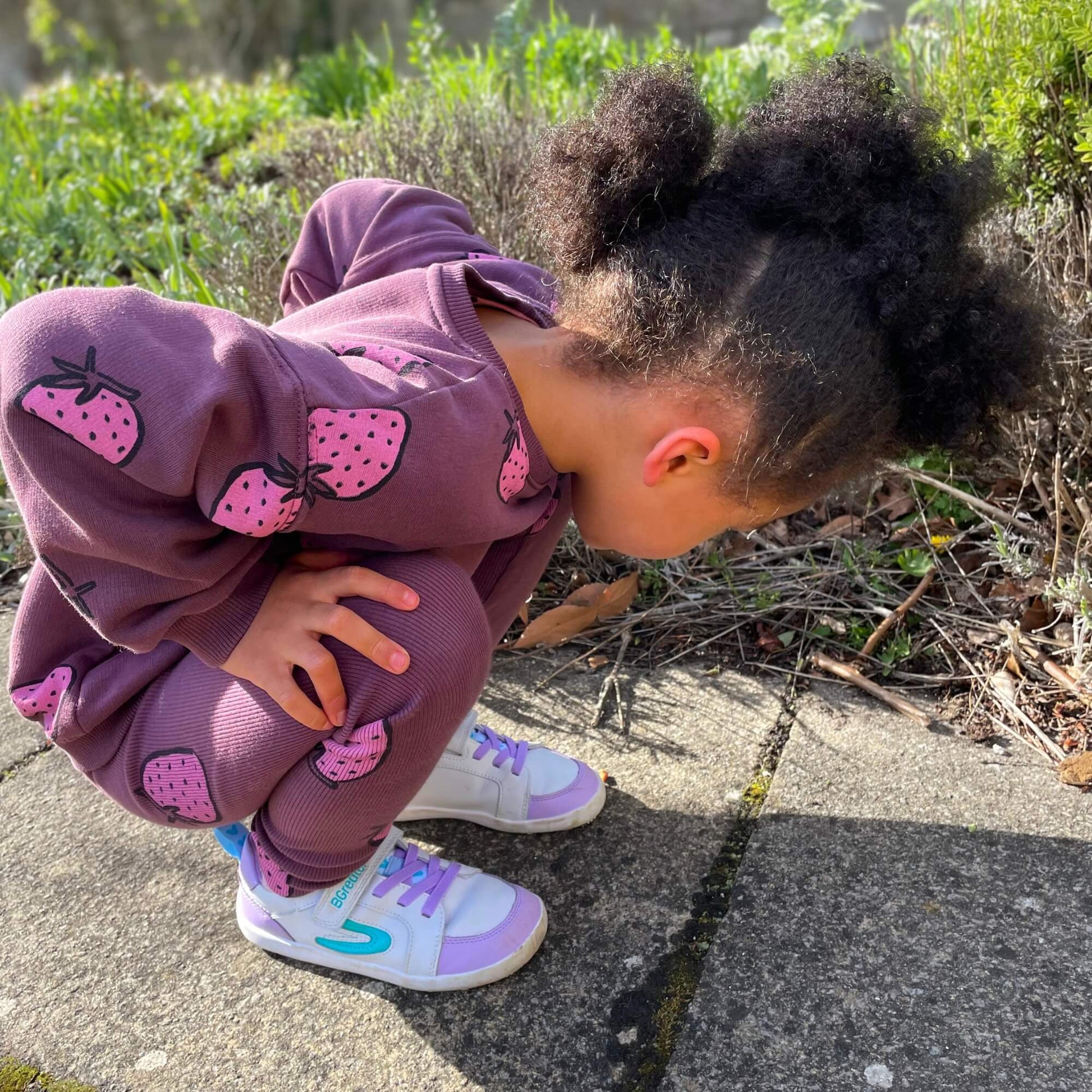 Kid wearing white and lilac leather barefoot trainers crouching on an outdoor pavement.