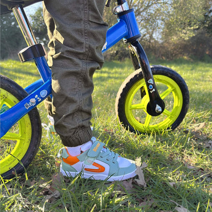 Kid's white, olive green and orange barefoot trainers shown outdoors next to a bike.