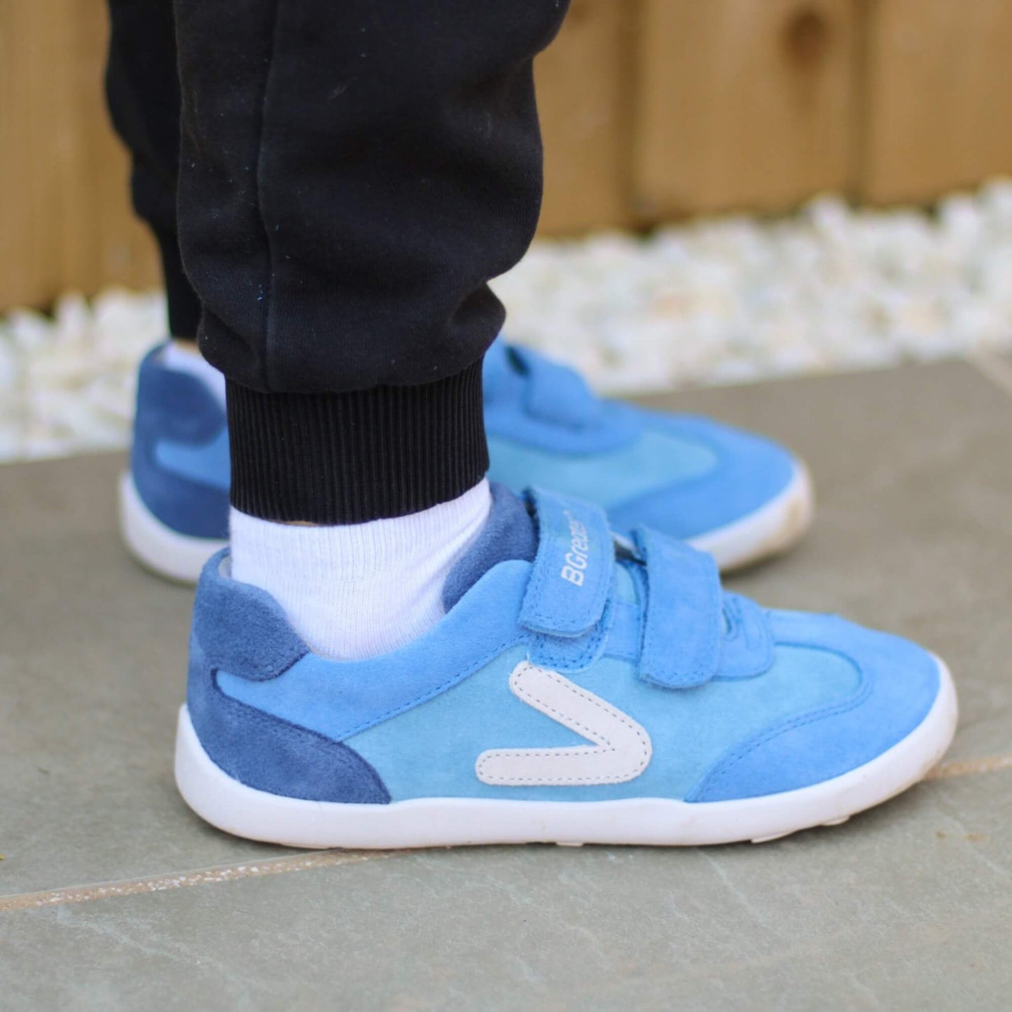 Kid wearing blue leather barefoot trainers with hook-and-loop straps on an outdoor stone patio.