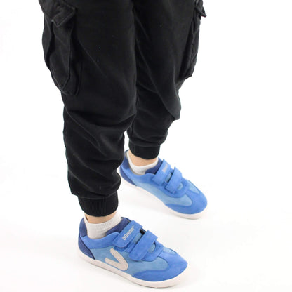 Kid sitting on a white background wearing blue leather barefoot trainers with hook-and-loop straps.