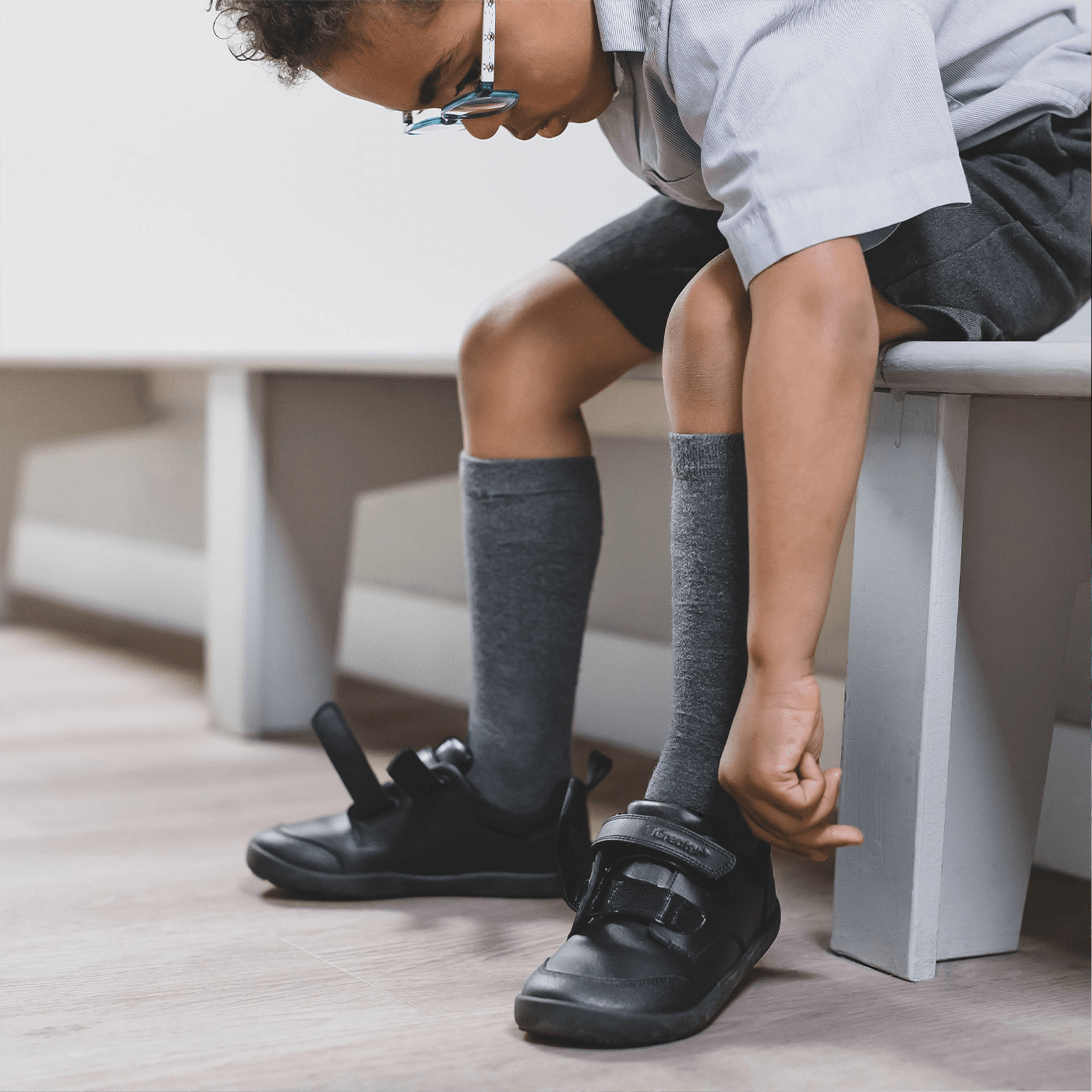 Boy sitting on an indoor bench putting on black leather barefoot school shoes with hook-and-loop straps.
