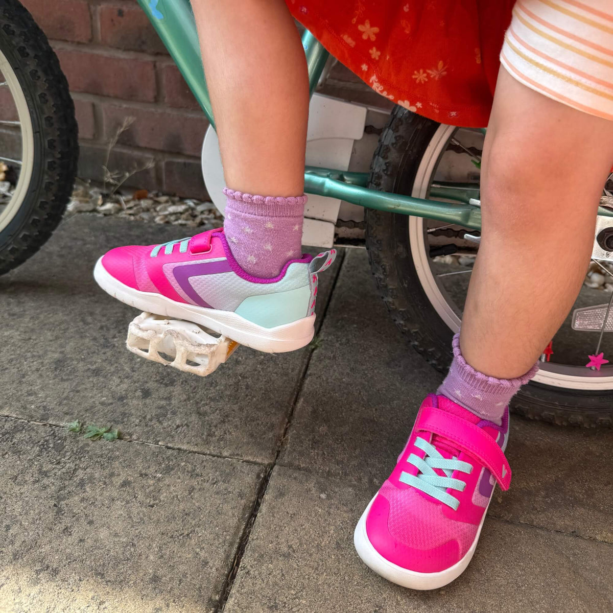 Close-up of kid's pink and teal barefoot trainers with elastic laces on a bicycle outdoors.